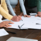 Focus sulle mani di persone, appoggiate a un tavolo, che consultano tutta una serie di documenti